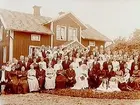 En och en halvvånings bostadshus med takhuv och förstubro i snickarglädje.
Bröllop. Brudpar och bröllopsgäster framför huset.
Aug. Andersson