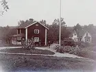 Tvåvånings bostadshus med glasad veranda. 
En och en halvvånings villa med burspråk och förstebro. En man med vattenslang.
Grosshandlare Uno Bergvall