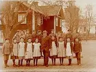 Tysslinge kyrkskola, tvåvånings vinkelbyggd skolbyggnad, 18 skolbarn med lärare Axel Holmberg.
Tysslinge kyrka i bakgrunden.