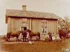 En och en halvvånings stuga, fyra äldre kvinnor framför stugan och en man med två hästar.