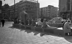 Semester i stan, 10 juli 1965

Under en solig dag på Våghustorget i Örebro sitter ett antal damer och herrar på en långbänk. Pumpen syns till vänster i bilden. Andra personer och bilar passerar.
