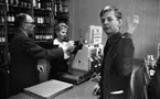 Stoppljus på systemet, 3 juni 1965

En yngre man handlar på Systembolaget. Han betjänas av en kassörska som står bakom disken. Hon håller ynglingens plånbok med identitetskort i handen. Bredvid kassörskan bakom disken står en man och betraktar tillsammans med henne den unge mannens legitimation. Han är klädd i mörk kostym och bär glasögon. I bakgrunden syns hyllor med vin- och ölflaskor. Även vid kassaapparaten står vinflaskor.
Kassörskan är klädd i mörka arbetskläder med vit krage upptill samt en vit blus inunder. Hon har små örhängen och kortklippt, fluffig frisyr. Ynglingen är klädd i mörk manchesterkostym samt mörk skjorta och slips. Han vinkar med ena handen mot kameran.