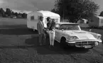 Camping 5 juli 1965.

Familj bestående av mor, far och liten flicka stående intill bil med husvagn på tältplats.