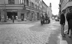 Hästar i stan, 8 juli 1965.

Man kör arbetshäst och vagn genom stan. Bredvid på trottoaren står en grupp unga män och samtalar.