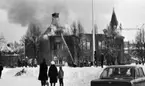 Noraskola brinner 18 februari 1966

Det brinner i en skola i Nora en vinterdag. I bildens bakgrund har brandsoldater riktat en brandspruta mot byggnadens tak för att försöka släcka elden med vatten. Grupper med skolelever står i snön på skolområdet och tittar på. Till höger i bakgrunden av bilden syns en kyrka och en lyftkran. I förgrunden av bilden står en bil med ett antal personer i. Till vänster syns en cykel som ligger i snön.