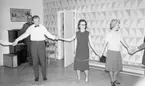 Dansskola, 15 februari 1966

Tre personer syns på bilden dansa i ring. Ytterligare personer dansar med dem men syns ej i helfigur. En man i bakgrunden spelar dragspel och har ett notställ framför sig.  Bakom honom står ett piano, en stol samt en resväska. På golvet syns det ett mönster av kontrasterande ljusare och mörkare kvadratiska rutor. Väggen har en mönstrad tapet.