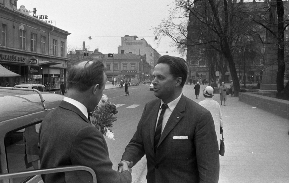 Trafikblomman, 5 maj 1966

Äldre man blir hyllad med blomsterbukett av man som skakar den äldre mannens hand, de står utanför en bil i stadsmiljö