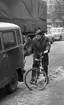 Dragos cyklar 28 januari 1966

En man sitter på en cykel. Han har satt ned vänsterfoten i marken och väntar vid ett stoppljus. Han är klädd i rock och byxor. Invid honom står en bil samt en delvis skymd man och i bakgrunden står en lastbil. Bakom denna står två ytterligare bilar på kö.