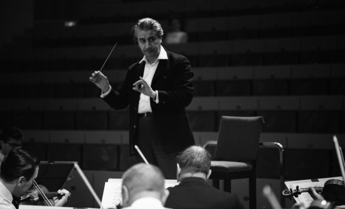 Cellibedache 13 juni 1966

En dirigent klädd i vit skjorta och svart kavaj viftar med taktpinnen framför en orkester. Framför honom sitter musiker och spelar.