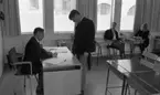 Beg. bilar, Ferieskola, Bodenkille slog rekord 14 juli 1966

En lärare sitter vid sin kateder och skriver på ett papper i ett klassrum. Han är klädd i mörk kostym, mörk slips samt vit skjorta. Framför honom står en ung man och betraktar vad läraren skriver på papperet. I bakgrunden sitter två andra unga män samt en ung kvinna vid varsin skolbänk. De befinner sig på ferieskola.