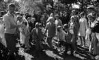 Midsommar, 26 juni 1967

Närbild på barn, kvinnor och män som dansar ringdans under midsommarfirande. Det är flera stycken ringar av dansande människor. En kvinna bär en vit kappa. En man till vänster håller i ett mikrofonstativ och har en cigarrett i munnen. En kvinna till höger är klädd i folkdräkt. Många män är klädda i kostymer.