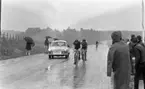 Orubricerad 18 maj 1967

Under en cykeltävling kommer tre tävlingscyklister cyklande på en väg klädda i idrottskläder och med hjälmar på huvudet. En bil kör också på vägen med en skylt på sig där det står: 