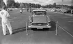 Orubricerad 21 augusti 1967

En man och en kvinna sitter i en Opel på en parkering. De är i färd med att köra iväg. En man i vit arbetsoverall står och pratar med en person till vänster. Ytterligare personer och bilar syns i bakgrunden. Även ett hus syns längre bort.