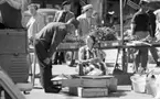 Kvar i stan 26 juli 1967

En kvinna i en kort ärmlös klänning sitter ned nära några lådor på ett torg i centrala Örebro. En kaffetermos står framför henne. En man står invid henne och böjer sig fram och talar med henne. Han är klädd i långärmad mörk tröja, mörka byxor, mörka strumpor och svarta skor. I bakgrunden står en bilar parkerade.