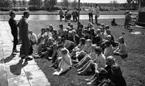 Glass, Nygren i Sten..., Gustavsvik 13 juni 1967

En grupp barn i tioårsåldern sitter på en gräsmatta i utomhusbadet Gustavsvik och lyssnar på en man klädd i ljus träningsoverall, mörka träskor och svarta solglasögon  som pratar till dem. En kvinna i mörk träningsoverll och vita träskor står bredvid honom. Andra personer syns i bakgrunden.