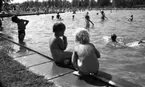 Grybe golf, Gustavsvik 17 juni 1967

Två små nakna barn befinner sig på utomhusbadet i Gustavsvik. Barnet till vänster sitter på bassängkanten och barnet till höger sitter på huk. I bakgrunden syns människor i alla åldrar som badar i bassängen. Många kvinnor och flickor badar i baddräkter och många män och pojkar badar i badbyxor. En barnvagn syns längre bort på marken på andra sidan bassängen.