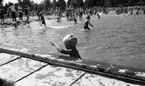 Grybe golf, Gustavsvik 17 juni 1967

Ett antal barn och vuxna badar i utomhusbadet på Gustavsvik. Många av flickorna och kvinnorna har baddräkter på sig och pojkarna är klädda i badbyxor. I förgrunden står en liten pojke nära bassängkanten i vattnet. Han lyfter sin vänstra arm.