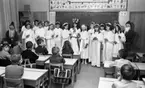 Lucia, Trafikolycka, Sörbyvägen Lillån 13 december 1966

Ett Luciatåg bestående av barn i tioåårsåldern står uppställt i ett klassrum i en skola. En Lucia befinner sig i mitten klädd i vitt linne, rött skärp om livet och ljuskrona med glitter på huvudet. Tärnorna som står runtomkring bär också vita linnen med glitter runt liven. De bär ljus i sina händer.Två tomtar står på ömse sidor om tärnorna. Framför Luciatåget sitter en publik i form av en skolklass.