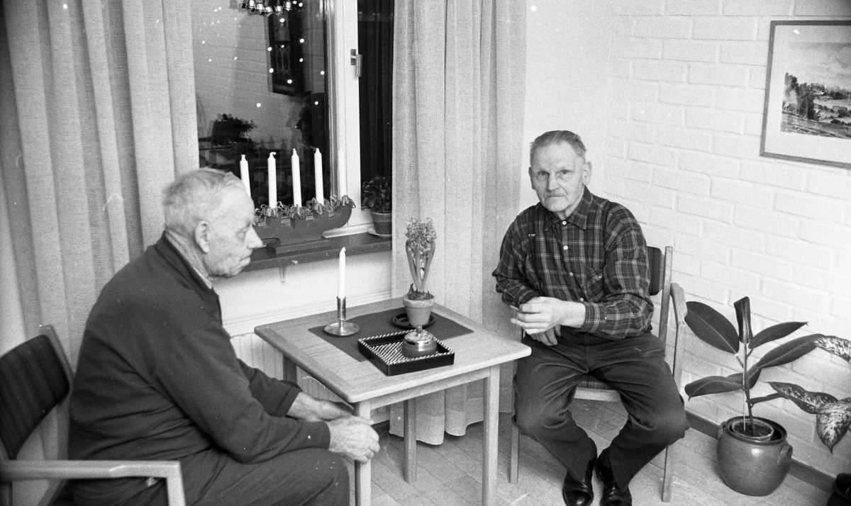 Ålderdomshotellet i Fjugesta 15 januari 1968

Två äldre män sitter vid ett bord på ett ålderdomshem i Fjugesta. Den ene mannen röker. Han har en cigarett i vänsterhanden. En askkopp står på bordet. En adventsljusstake står i fänstret i bakgrunden. En tavla hänger på väggen till höger.