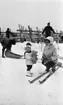 Ånnaboda 12 februari 1968

En liten flicka står i Ånnabodas skidbacke med en nalle i famnen. Hon är klädd i vit mössa med mörk tofs på huvudet, mörk halsduk, vit jacka, mörka byxor och vita stövlar. Invid henne knäböjer en kvinna klädd i vit mössa, vitmönstrad jacka, svarta byxor och svarta pjäxor. Hon har skidor på fötterna och håller ett par stavar klämda mot vänstra sidan av kroppen med vänster arm. Fler personer samt ett ställ med skidor syns i bakgrunden. En person lastar något i en pulka.