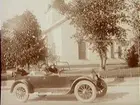 Exteriör. Bil med Ellen Larsson vid ratten, herr och fru Larsson som medresenärer. I bakgrunden Lindesbergs kyrka. 
Bilen är en Oldsmobile modell 30 från 1923-24 med en 42 hästkrafters sexcylindrig motor. Den registrerades den 2:e april 1924 på fröken Jenny Larsson, Örebro, och fick registreringsnummer T99. År 1930 ägdes den fortfarande av Jenny Larsson.