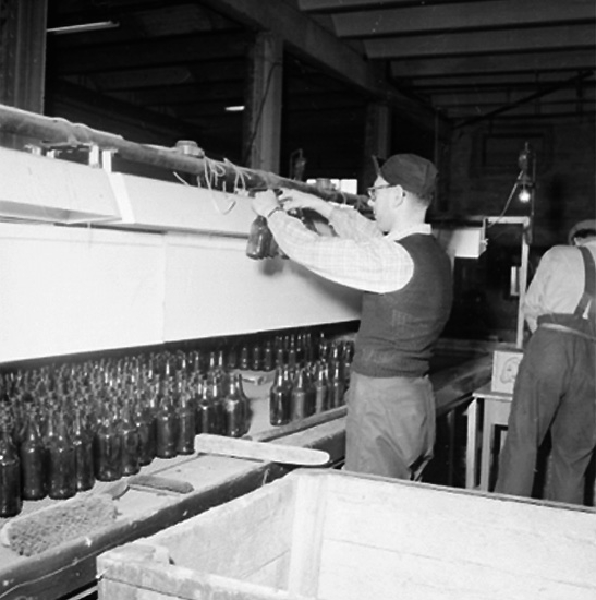 Hammars glasbruk, interiör, två män.
Mannen längs fram är Isak Karlsson f, född 1917-10-22 och död 1988-05-29. Han anställdes vid Hammars Glasbruk 1956-04-19. På bilden står han vid kylröret och synar glasflaskorna för ev skador och fel, bl a "tuppapinne".