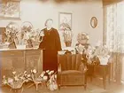 Rumsinteriör, en kvinna.
Fru Hellmér omgiven av blommor (högtidsdag).