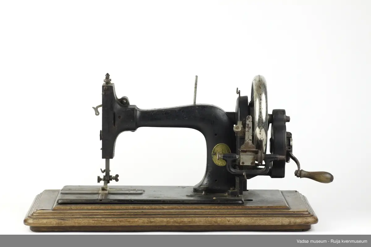 Hånddrevet symaskin av typen Model B, produsert i 1896 av G.M. Pfaff Kaiserslautern, Tyskland. Maskinen er hengslet fast i en profilert sokkel i treverk. Inni sokkelen finnes et rom beregnet til diverse symateriell. Rommet nås gjennom å løfte maskinen fra brukersiden. I sokkelen, parallelt med maskinens lengderetning, er det innfelt en målestokk.