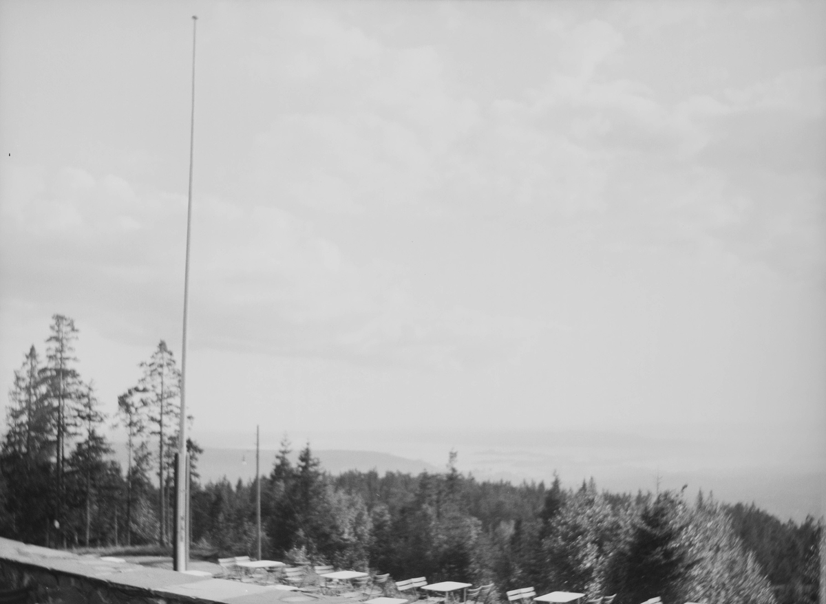 Utsikt fra Grefsenkollen restaurant i Oslo. I forgrunnen sees en mur og nederste terasse hvor det er plassert bord og stoler. Ellers ser man utover skogen. Man skimter Oslofjorden mellom skog og himmel.