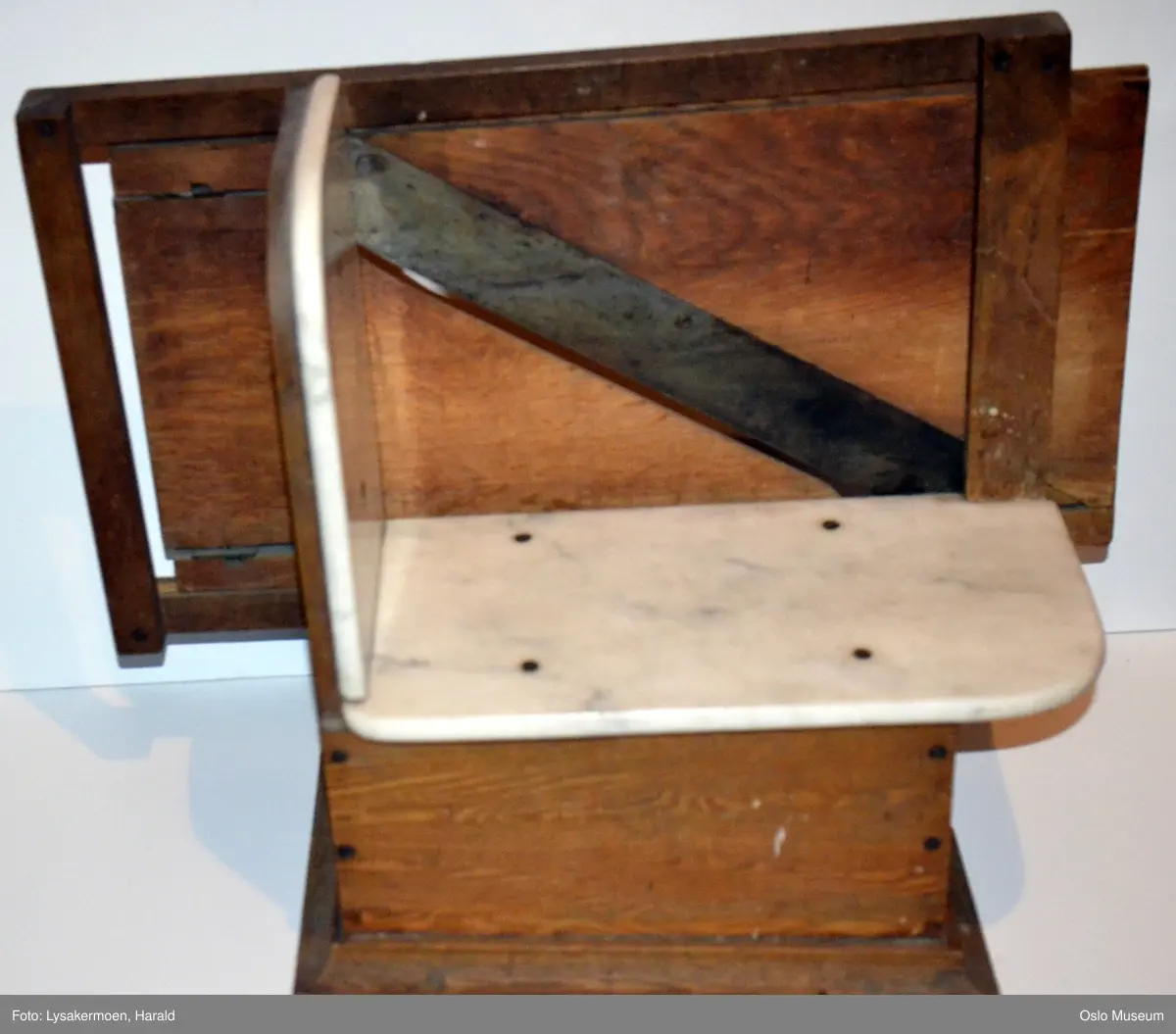 En profilert list som ramme om en glatt fotkasse. På toppen av kassen hviler en marmorplate, en annen marmorplate står perpendikulært på denne. På baksiden er en ramme hvor hele brettet med knivene kan beveges fram og tilbake. Brettet har håndtak og en skruanordning.