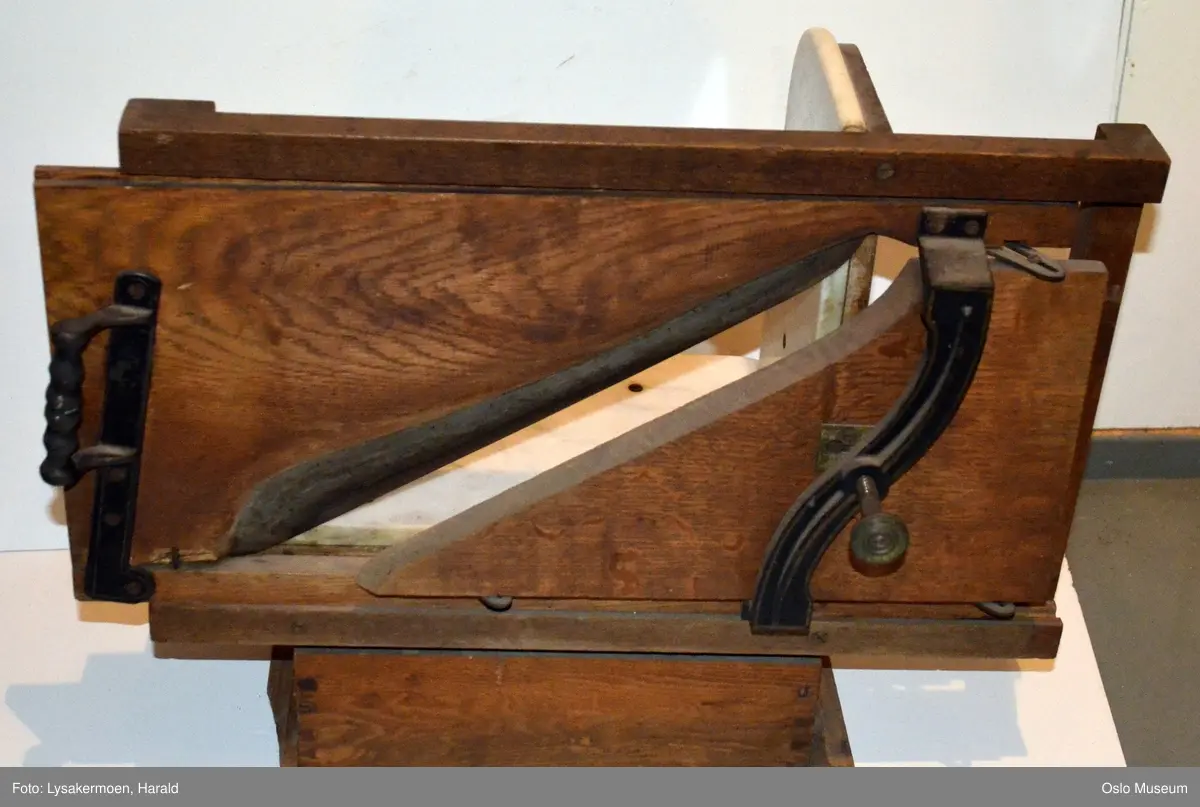 En profilert list som ramme om en glatt fotkasse. På toppen av kassen hviler en marmorplate, en annen marmorplate står perpendikulært på denne. På baksiden er en ramme hvor hele brettet med knivene kan beveges fram og tilbake. Brettet har håndtak og en skruanordning.