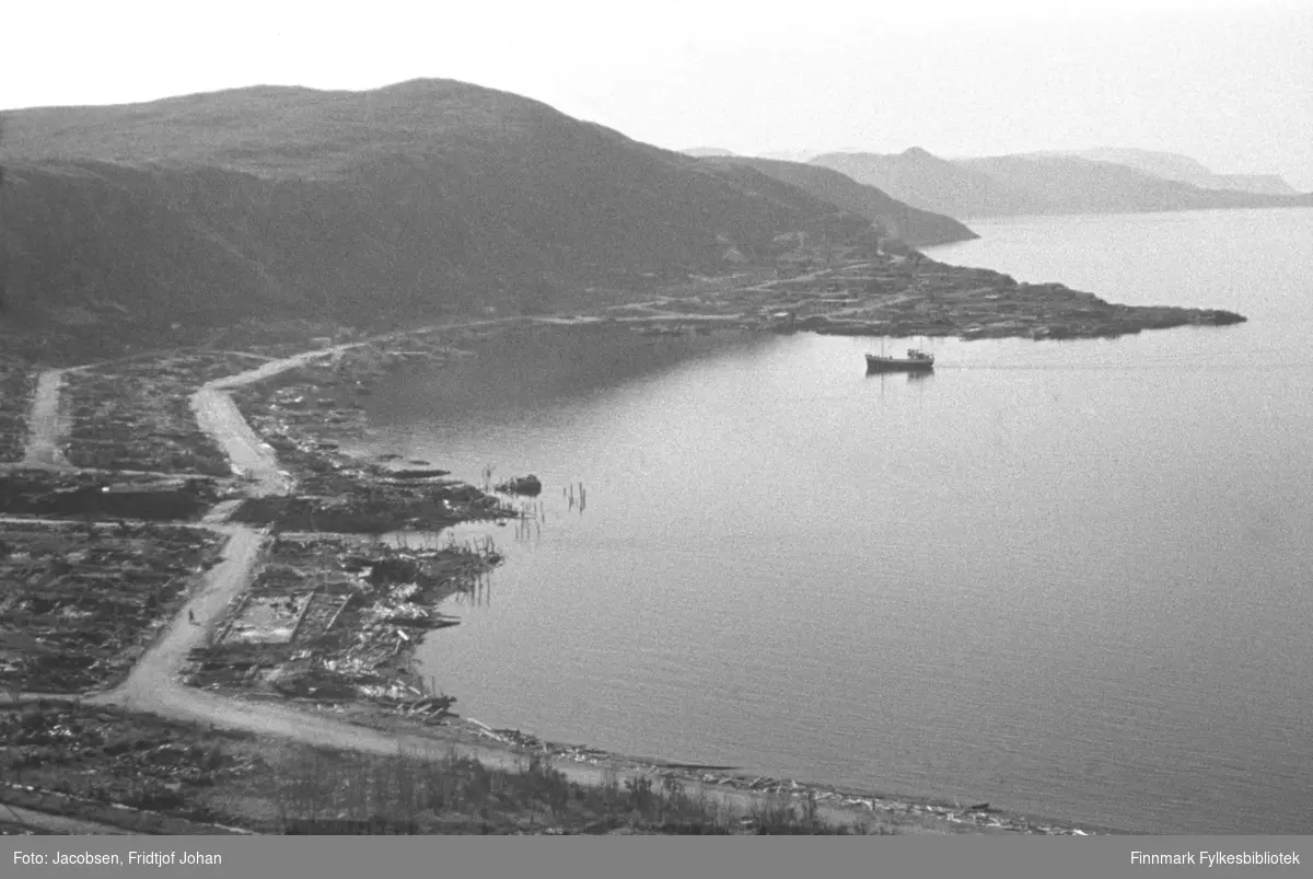 Et totalt rasert Hammerfest ses fra Mollafjell. Noen gresstuster vises i skråningen ned fra Mollafjell. Bydelen Molla ses nederst, adskilt fra sentrum ved Storelva som skjærer gjennom terrenget til venstre på bildet. Fuglnesveien går langs strandkanten fra Storelva og videre bortover mot Fuglenes. På andre siden av Storelva går Nedre Grønnevoldsgate, Strandgata i dag, gjennom sentrum og opp mot kirka. Veien ved foten av Fjellet salen til venstre på bildet er Elvebakkveien, som også går fra Storelva mot sentrum. En skøyte er på tur inn mot havna forbi Hammerfest-neset og rett over det ses Rypklubben. Øverst på neset er bydelen Haugen. Litt diffust ses øya Seiland i bakgrunnen.