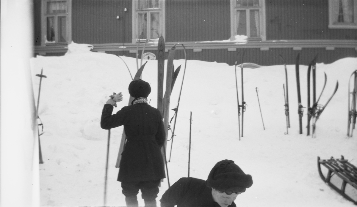 To personer holder på med skiutstyr utenfor en Haugastøl stasjon og turisthotell. Mange par ski og staver samt en kjelke står rundt omkring.