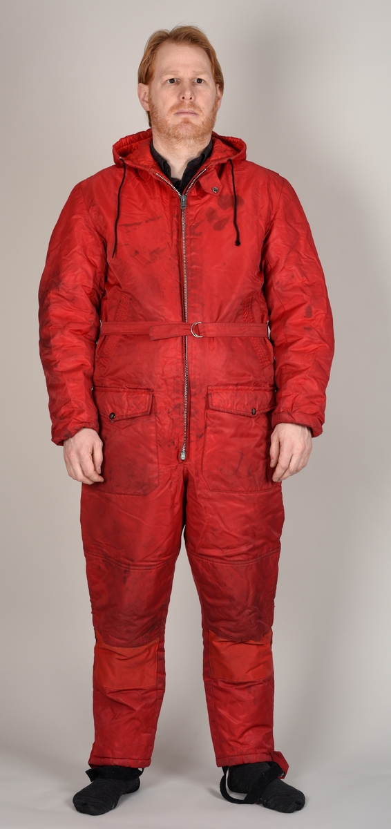 Rød dress med glidelås, hette, påsydd belte og to bukselommer.
Hetten har glidelås på oversiden.
Det er sydd på en sort stropp under hvert bein for å feste under skoene. Beltet lukkes med to halvrunde metallhemper.