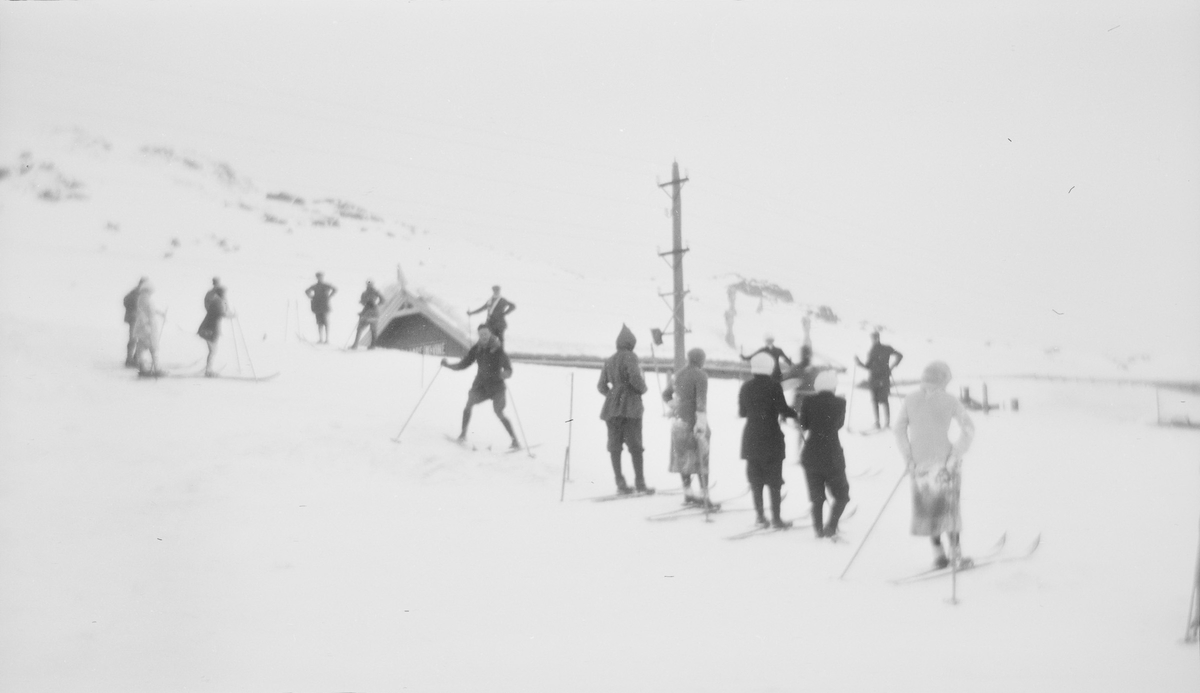 Menn og kvinner står på hver sin side av en bakke eller løype, de fleste med ski på bena. I bakgrunnen sees nedsnødde bygninger, fjell og en overbygget del av jernbanen.