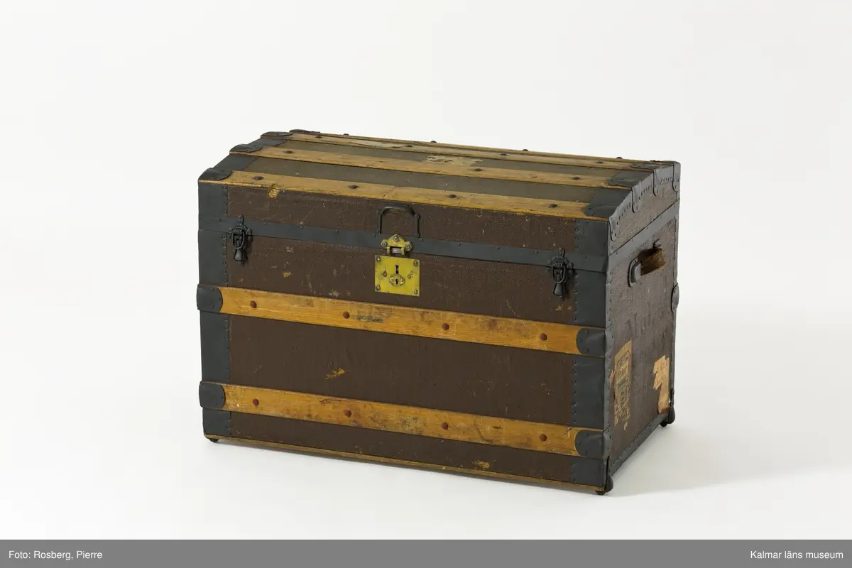 KLM 39675. Koffert, reskoffert, trä, papp, textil, läder, metall. Koffert för förvaring och resor, av trä, klädd med väv, beslag av trä och järn, lås av metall, två spännen och ett handtag av läder på varje gavel. På locket fyra beslag av trä och två på fronten. Insidan är klädd med papp eller tapet med rutigt mönster i rött, grönt och blått på gul botten. 
På lockets insida en fastklistrad tillverkaretikett med rubriken: NATHAN NEAT & CO TRUNKS and Travelling Equipage (för hela texten se foto). Text i svart på höger gavel: M.J. Boston Mass. Spår av gamla resetiketter på höger gavel. Kofferten är brunmålad. Inköpt i Amerika.