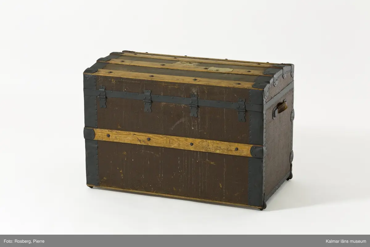 KLM 39675. Koffert, reskoffert, trä, papp, textil, läder, metall. Koffert för förvaring och resor, av trä, klädd med väv, beslag av trä och järn, lås av metall, två spännen och ett handtag av läder på varje gavel. På locket fyra beslag av trä och två på fronten. Insidan är klädd med papp eller tapet med rutigt mönster i rött, grönt och blått på gul botten. 
På lockets insida en fastklistrad tillverkaretikett med rubriken: NATHAN NEAT & CO TRUNKS and Travelling Equipage (för hela texten se foto). Text i svart på höger gavel: M.J. Boston Mass. Spår av gamla resetiketter på höger gavel. Kofferten är brunmålad. Inköpt i Amerika.