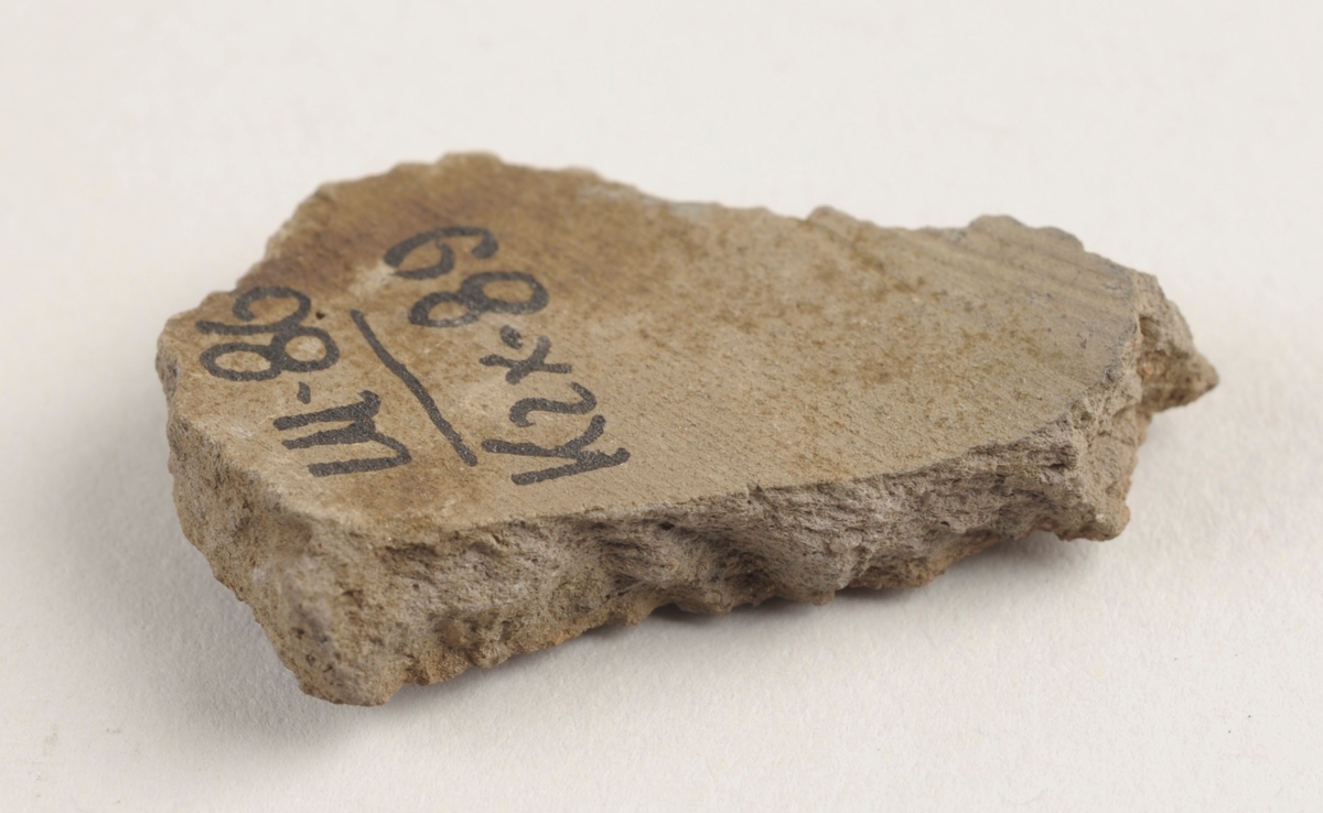 Flatt keramikkfragment med bruddkanter. Fragmentet er grått i farge.
