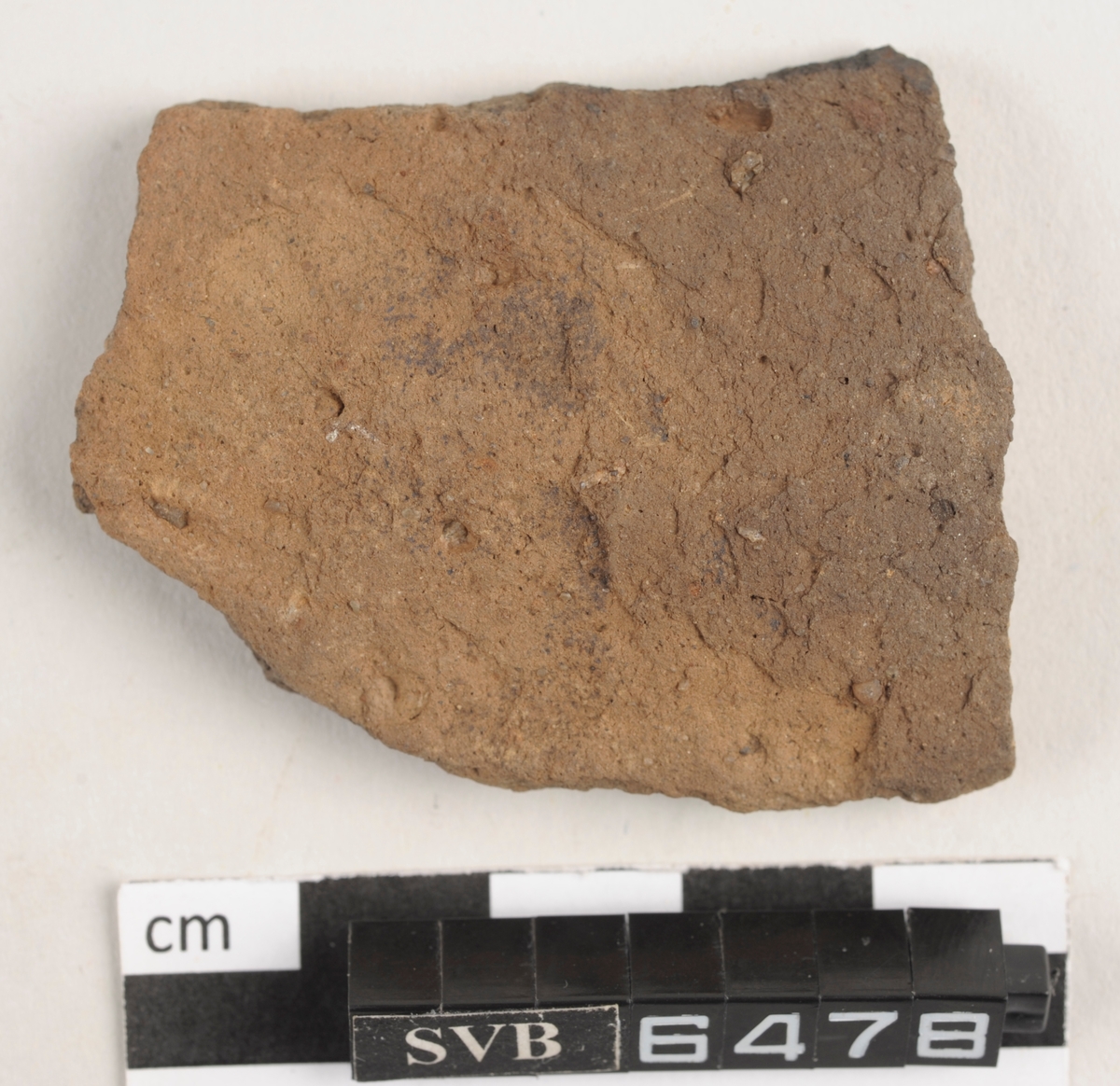 Lett buet keramikkfragment med bruddkanter. Fragmentet har en rødlig farge.