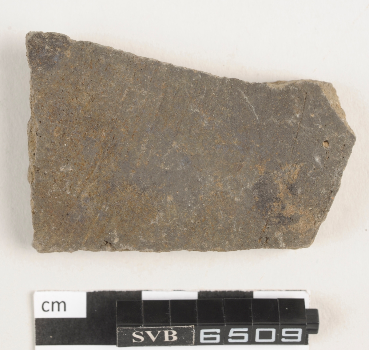 Flatt keramikkfragment med bruddkanter. Fragmentet er brunt i farge.
