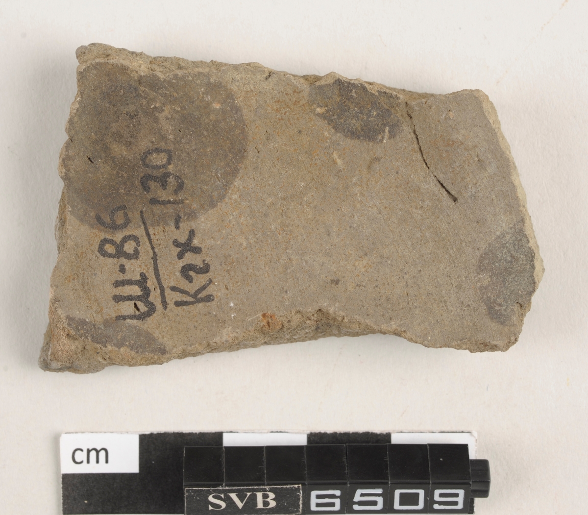 Flatt keramikkfragment med bruddkanter. Fragmentet er brunt i farge.