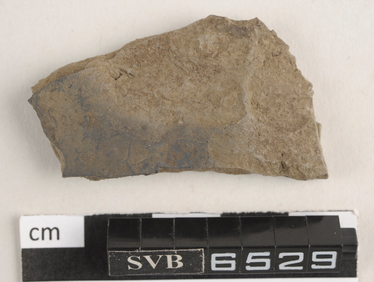 Flatt keramikkfragment med bruddkanter. Fragmentet er grått i farge.