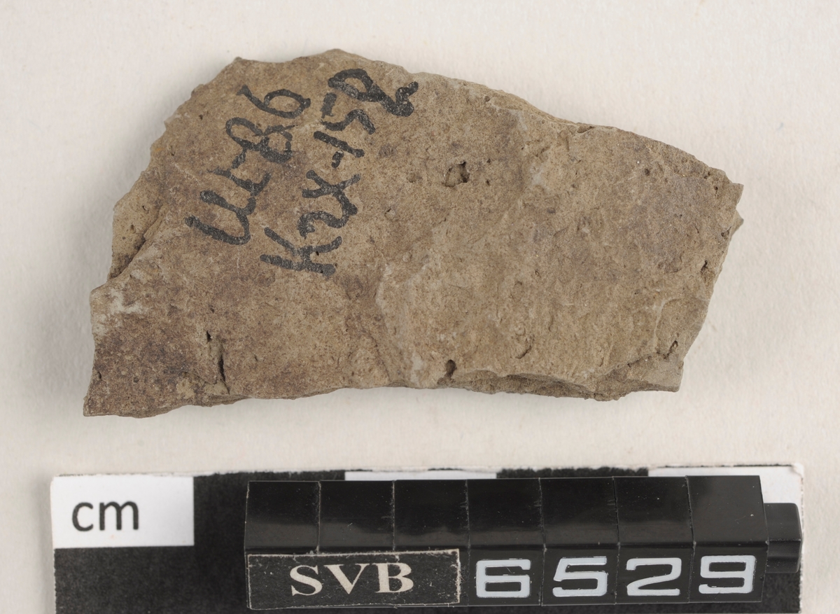 Flatt keramikkfragment med bruddkanter. Fragmentet er grått i farge.