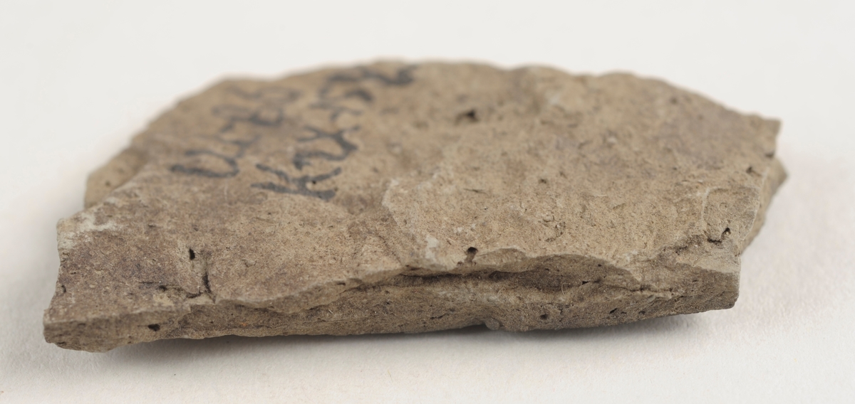 Flatt keramikkfragment med bruddkanter. Fragmentet er grått i farge.