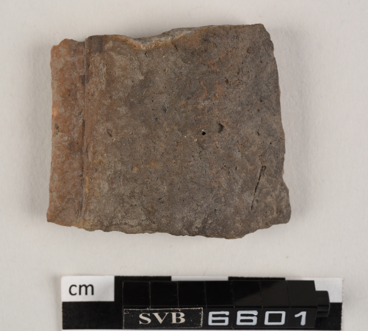 Buet keramikkfragment med bruddkanter. Fragmentet er grått i farge på utsiden og oransje i farge på innsiden.