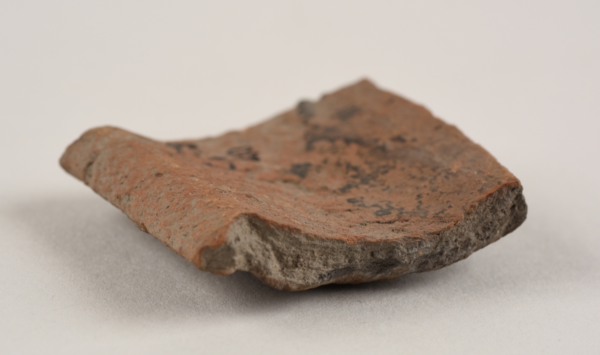 Buet keramikkfragment med bruddkanter. Fragmentet er grått i farge på utsiden og oransje i farge på innsiden.