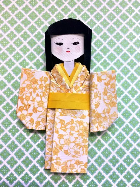 Papirdukke brettet i japanske origamiteknikk. Dukken har fått kimono i gult, blomstret papir.