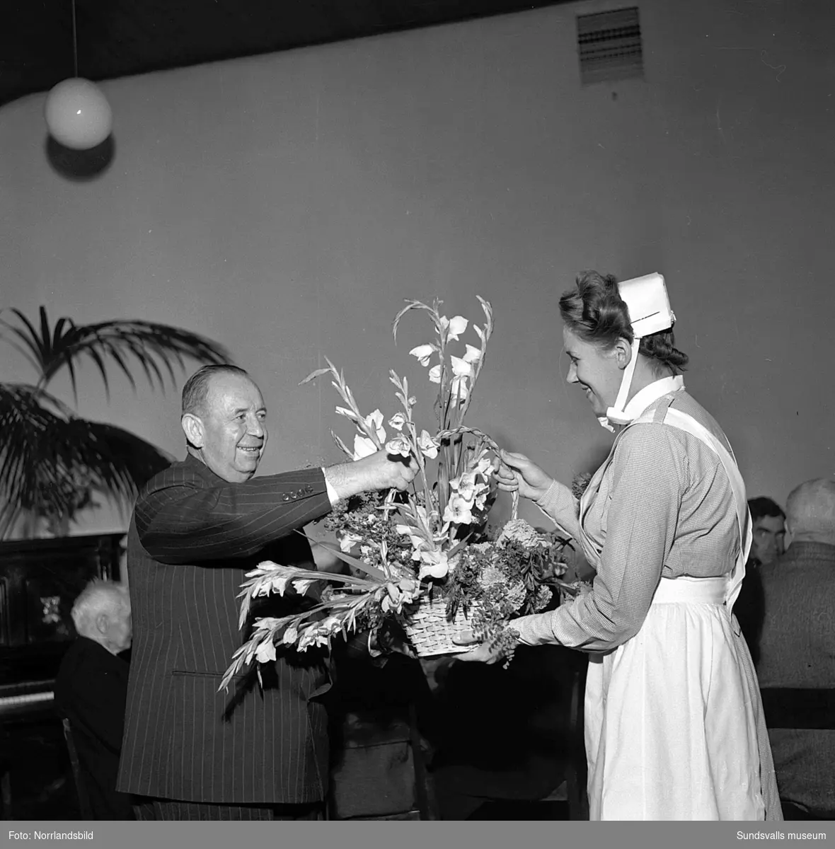 Lindgrens järnhandel firar 100-årsjubileum. Direktör Fridolf Brunnzell uppvaktar fru Palmqvist med en minnesskrift, samt kaffekalas på ett ålderdomshem.