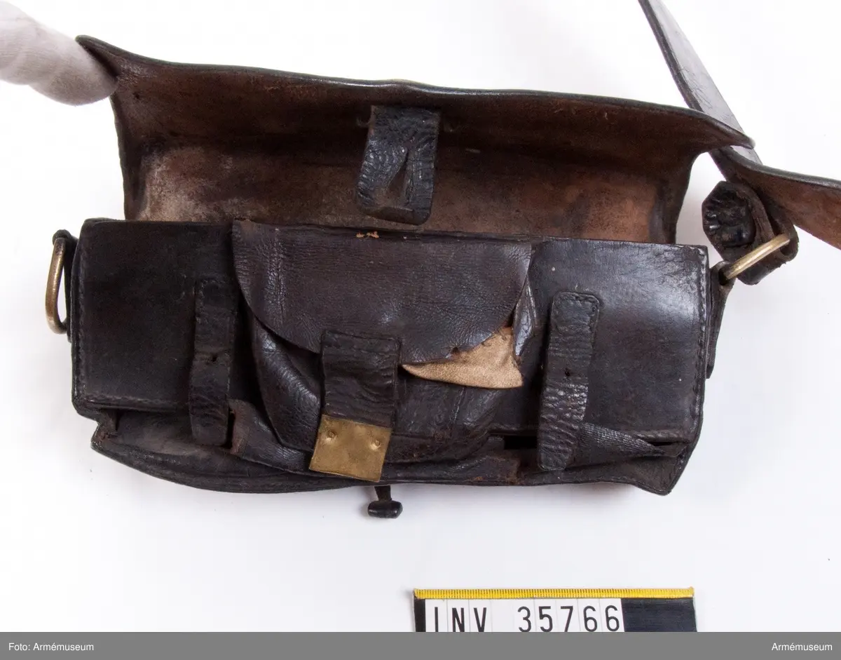 Kartuschväska av svart läder med mässingsbeslag i form av Skånes vapen. Kartuschen har en extra ficka för knallhattar och ett fack i botten som stängs med två remmar. De tillhörande spännena saknas. I kartuschen finns en träkloss med borrade hål med ca 18 mm diameter.