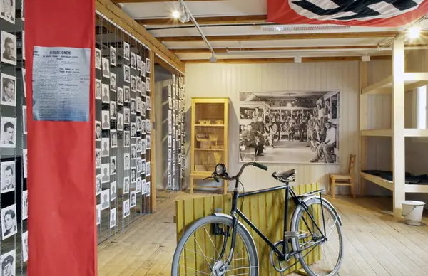 Bilde av utstillingen "Skudd i mørket - hva skjedde i Trandumskogen?" Rødt bånd med artikkel til venstre, sykkel på midten, brakkeseng til høyre, bilder av henrettede menn helt til venstre, stort bilde av krigsfanger og et monter med gjenstander bakers i rommet.